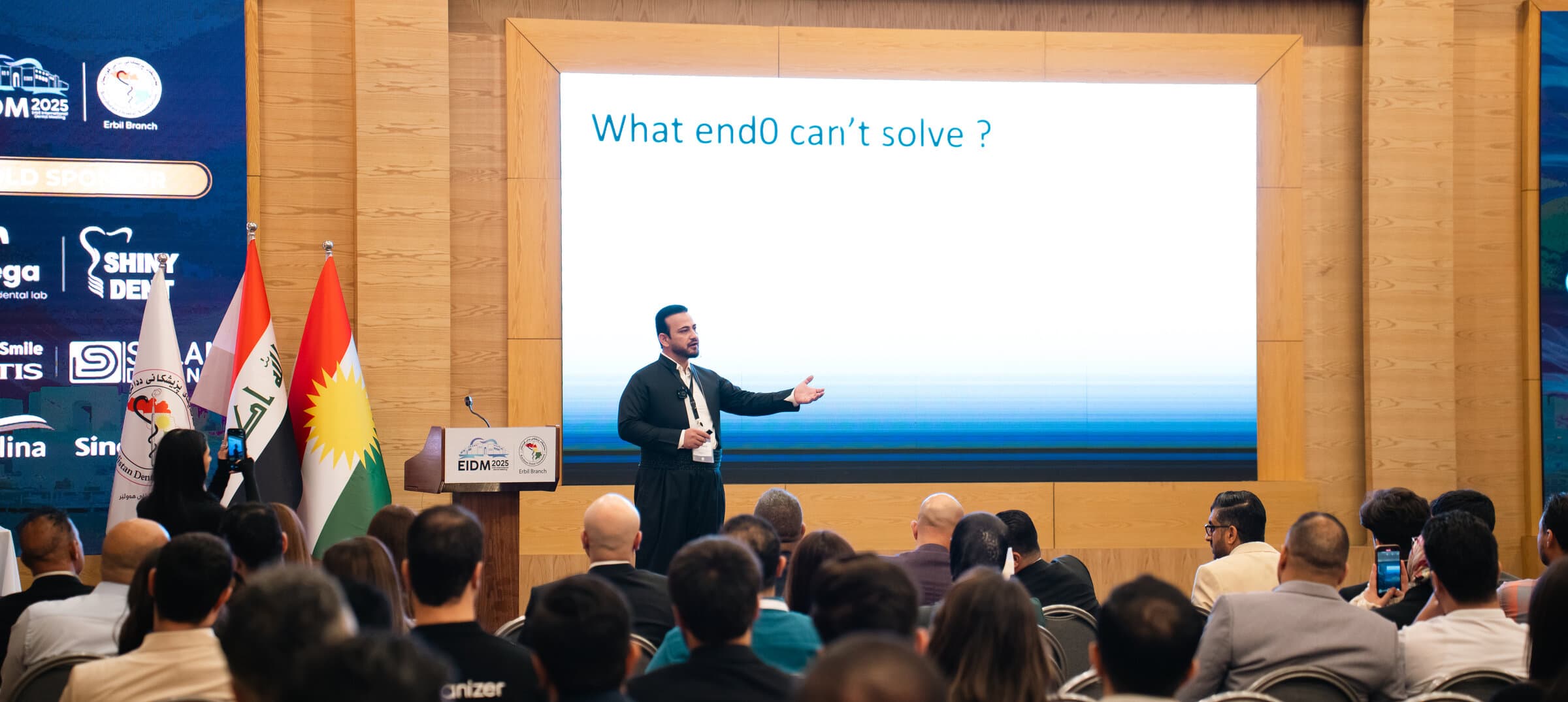 Dr. Jegr Koyi lecturing on endodontics to a packed hall at a dental conference, Kurdistan flag beside the stage